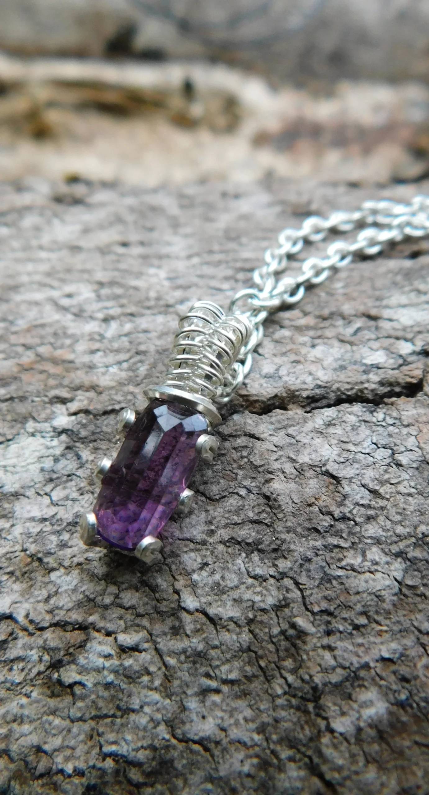 Close-up of a silver chain necklace with a purple gemstone pendant resting on a textured rock surface.